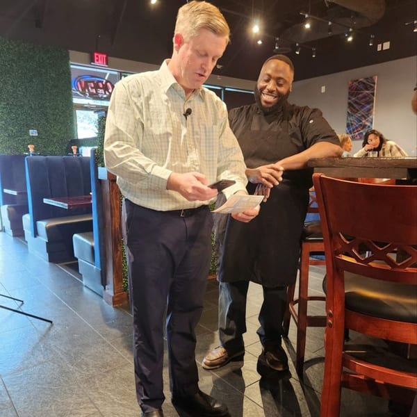 A man in a plaid shirt looks at a paper while a chef in a black apron smiles beside him.