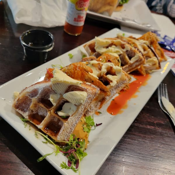 Chicken and waffles with syrup, powdered sugar, and butter, garnished with microgreens on a white plate.