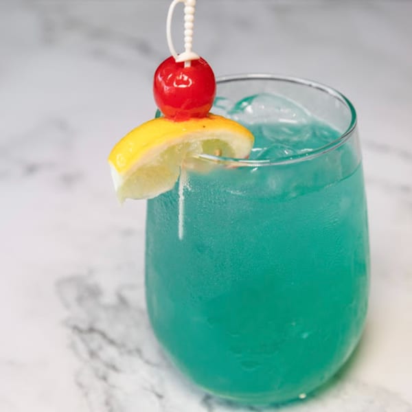 A vibrant blue cocktail with ice, garnished with a lemon slice and a red cherry on a white marble surface.