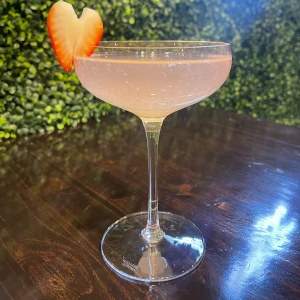 A light pink cocktail in a coupe glass, garnished with a heart-shaped strawberry slice.