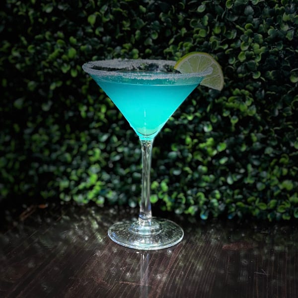 A vibrant blue cocktail in a martini glass with a sugared rim and lime wedge, against a green leafy background.