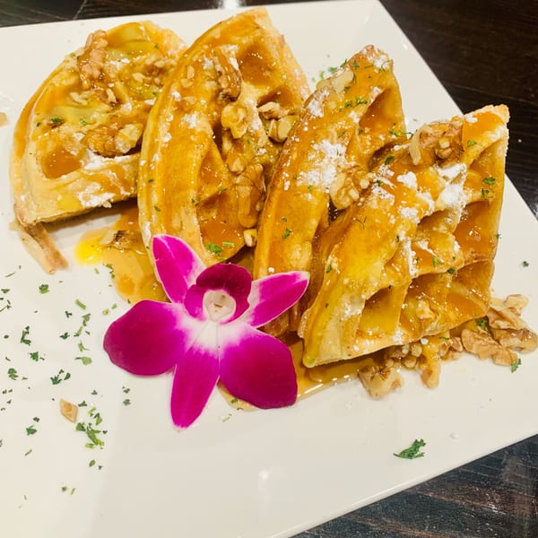 Golden waffles with syrup, walnuts, powdered sugar, and a pink orchid on a white plate.