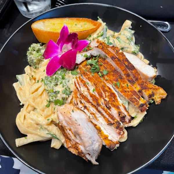 Blackened chicken pasta with broccoli, garlic bread, and an orchid garnish on a black plate.