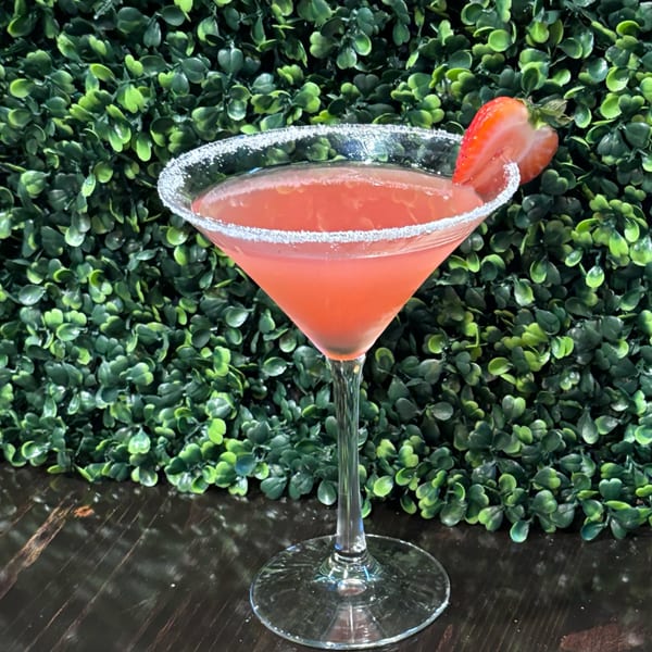 Pink cocktail in a martini glass with a sugared rim and strawberry garnish against a green leafy wall.