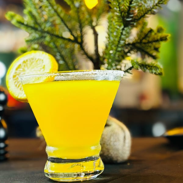 A vibrant yellow cocktail in a martini glass, garnished with a lemon slice, against a festive green background.