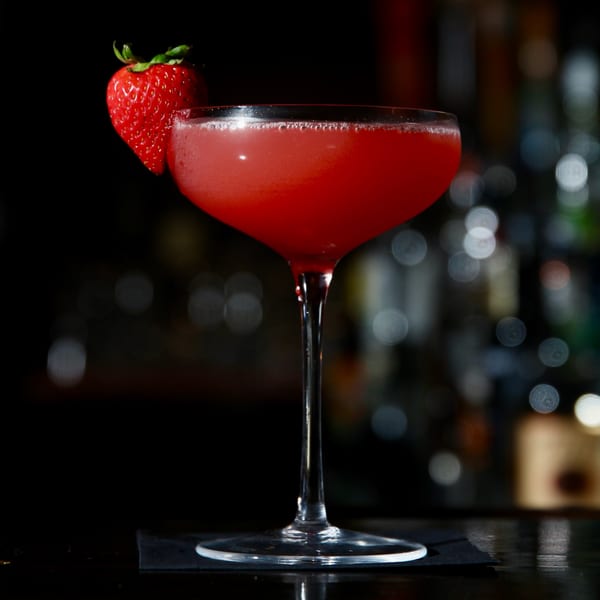 A vibrant red cocktail in a coupe glass, garnished with a fresh strawberry, against a dark, blurred background.